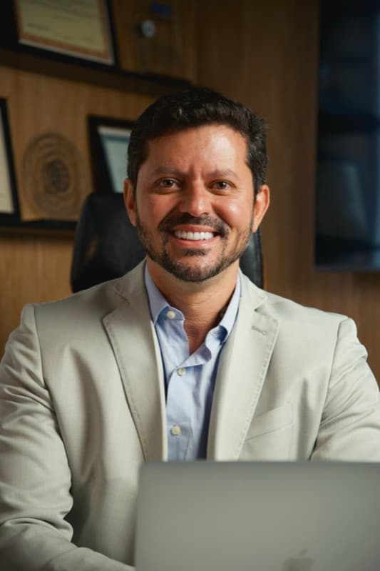 Dr. Flávio Pinheiro sorrindo à mesa do consultório, com seus diplomas na parede ao fundo