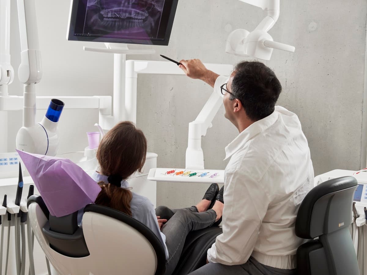 Dentista aponta para uma radiografia panorâmica na tela enquanto explica o tratamento à paciente na cadeira